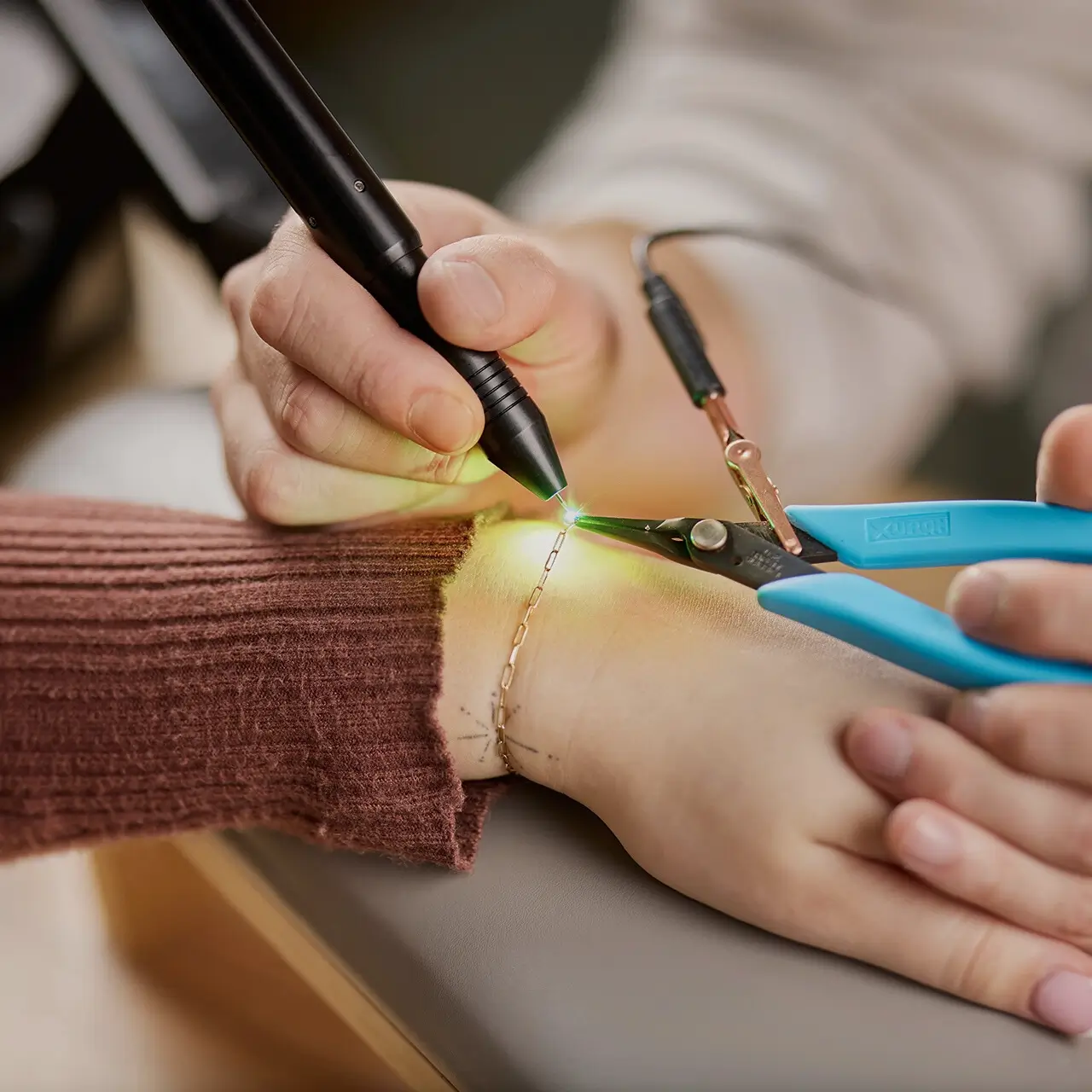 Close-up of a permanent jewellery welding process, showing a gold chain being expertly welded onto a wrist using a precision welding tool and blue pliers.