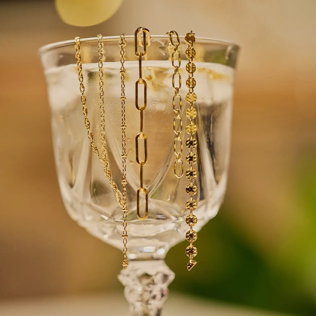 Close-up of waterproof gold permanent jewellery chains draped over a crystal glass, showcasing elegant designs with a soft, blurred background.